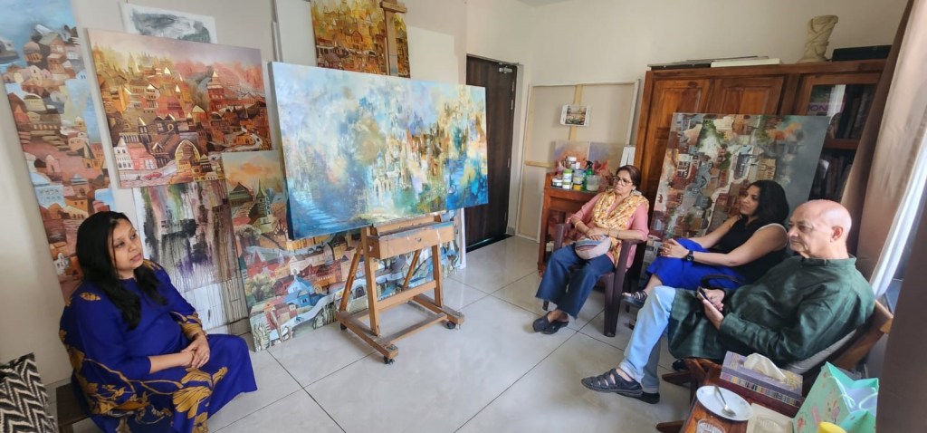 A group of four people seated in an art studio, surrounded by colorful paintings. One woman is sitting on the floor in a blue dress, while the others are seated in chairs, engaged in conversation. An easel with a partially completed painting is in the foreground.