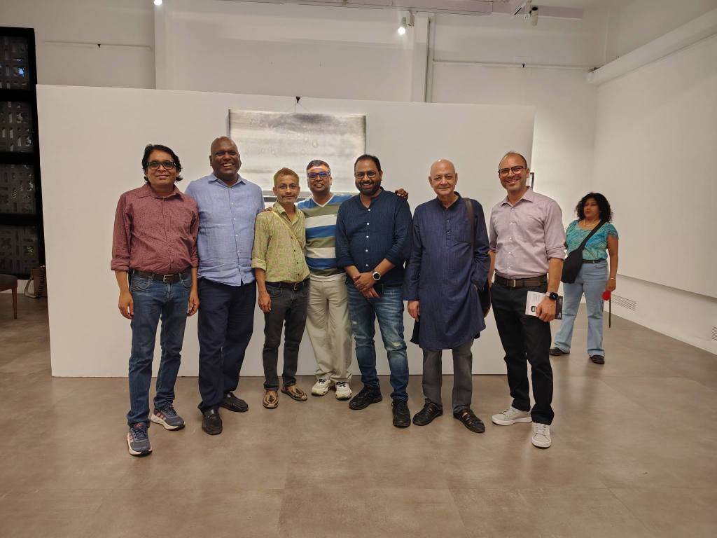 A group of seven men posing for a photo in an art gallery, standing in front of a large white wall with an abstract painting. Behind them, a woman is seen in the background, slightly out of focus.
