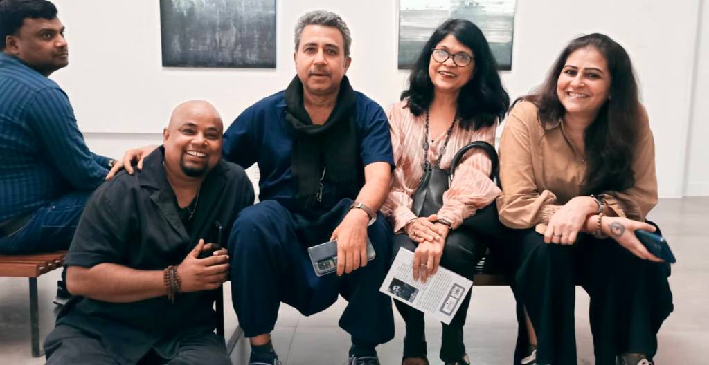 A group of four people seated in an art gallery, smiling at the camera. Two men are in the foreground, one of whom is holding a phone and the other is wearing a black shirt. Two women are seated next to them, one wearing a light blouse and the other with glasses, both holding bags. A man appears in the background.