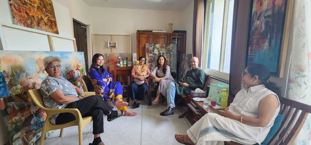 A group of six people sitting in a well-lit room, surrounded by artwork and creative materials. They appear engaged in conversation, with a mix of casual and traditional attire.
