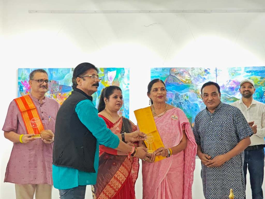 A group of five individuals interacting at an event, with some holding awards or trophies. The background features colorful paintings.