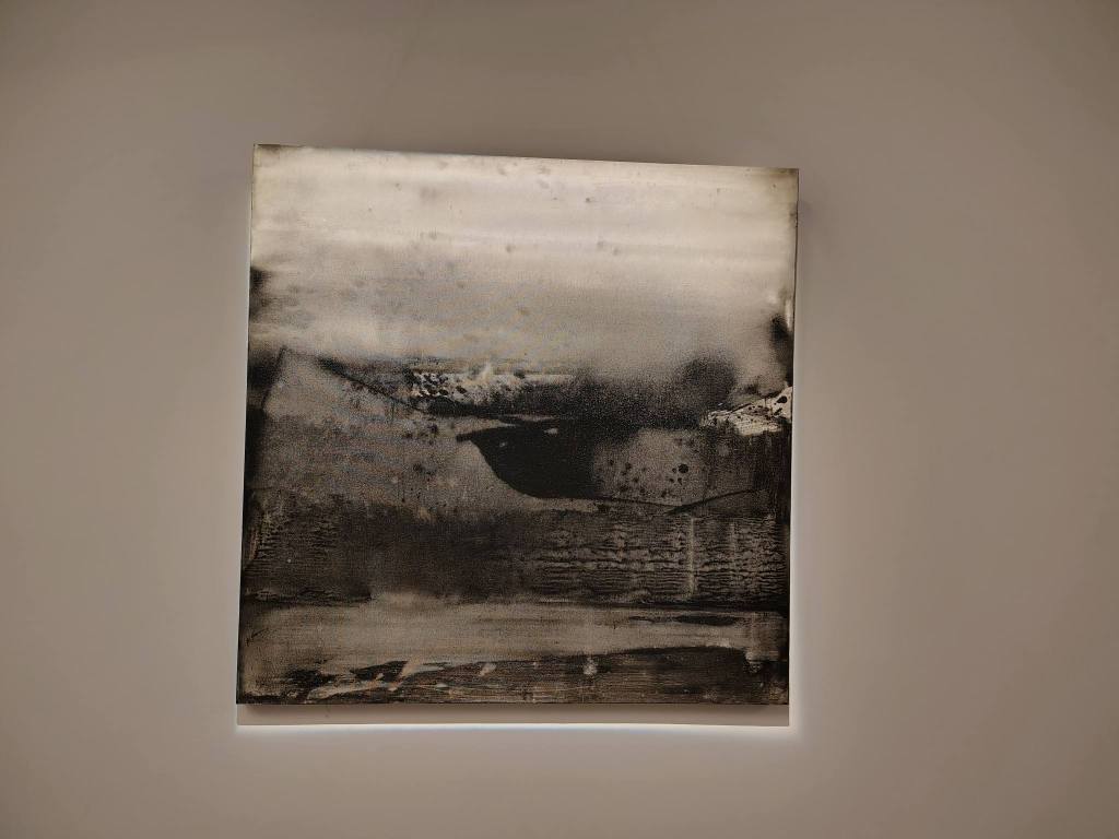 An abstract painting featuring shades of black and gray, depicting textured shapes and blurred forms, displayed on a wall with soft lighting.