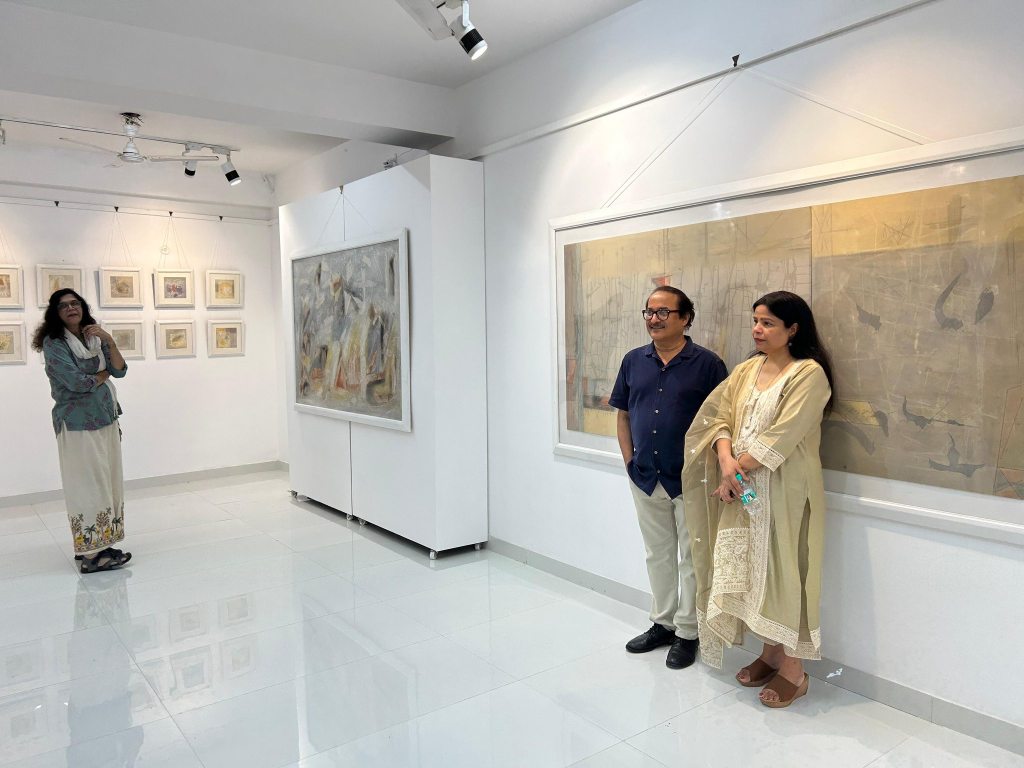 A group of three individuals standing in an art gallery, with paintings displayed on the walls. One woman appears in the background, while a man and a woman stand in the foreground next to a large artwork.