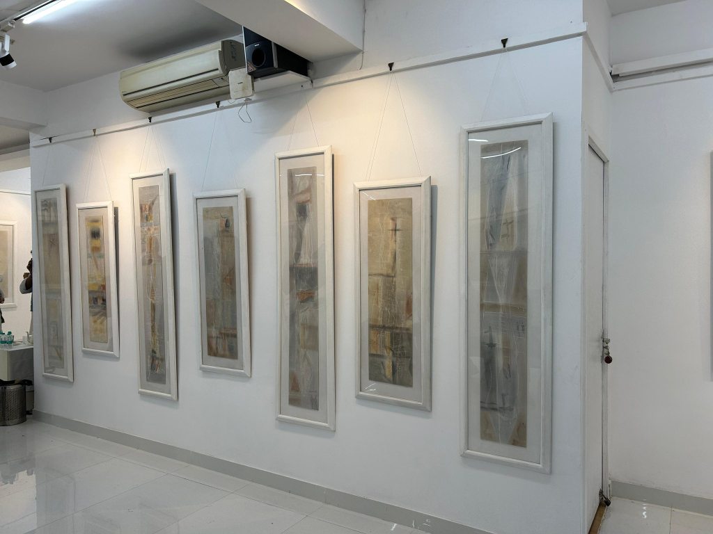 An art gallery displaying several tall, framed artworks hanging on a white wall, with a minimalist interior and polished floor.