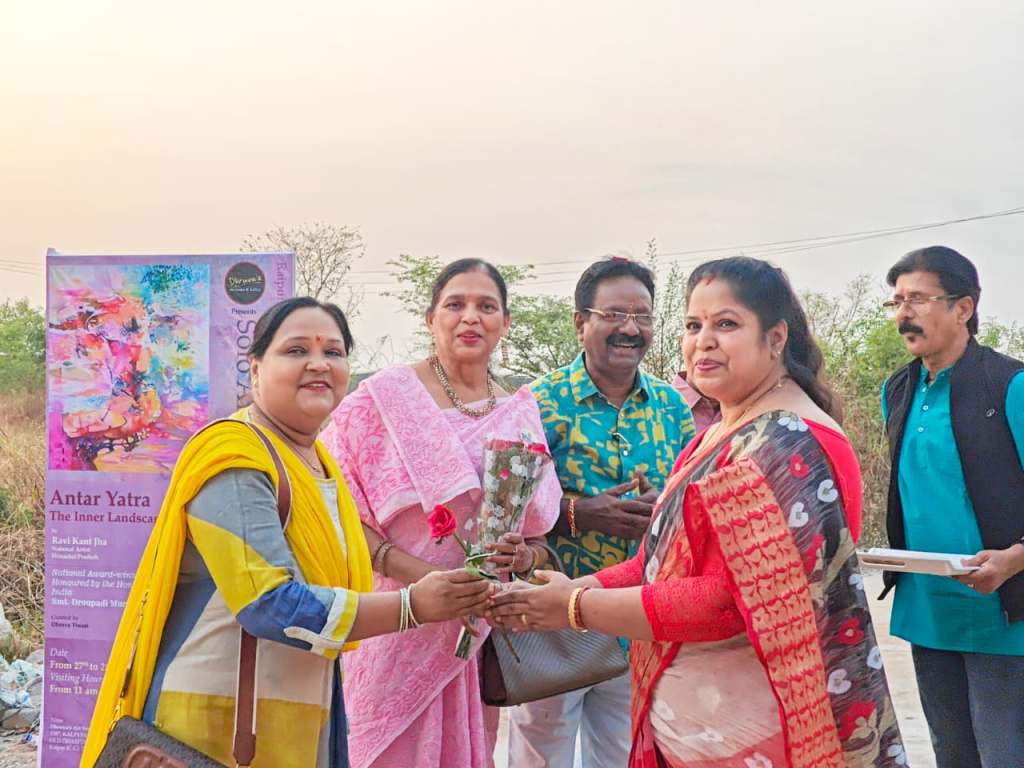 A group of five people stands outdoors with a banner behind them that reads 'Antar Yatra: The Inner Landscape.' Two women are exchanging flowers, while the others are smiling in the background.