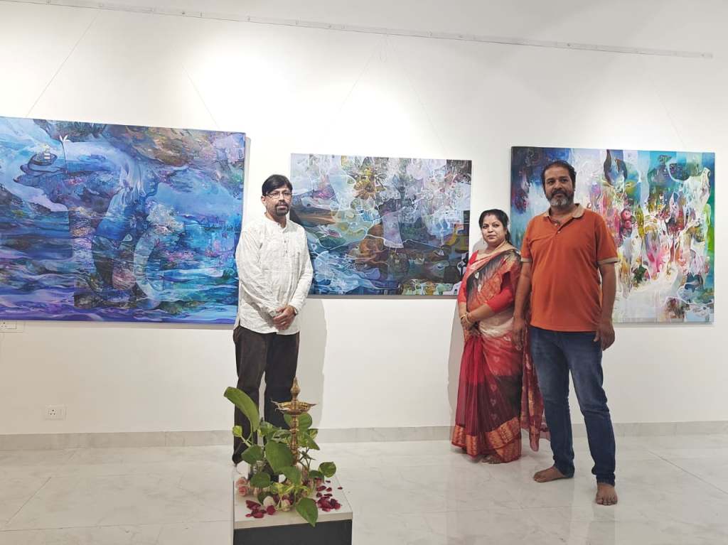 Three individuals posing in an art gallery with colorful abstract paintings on the walls. A decorative lamp is placed in the foreground, surrounded by flower petals.