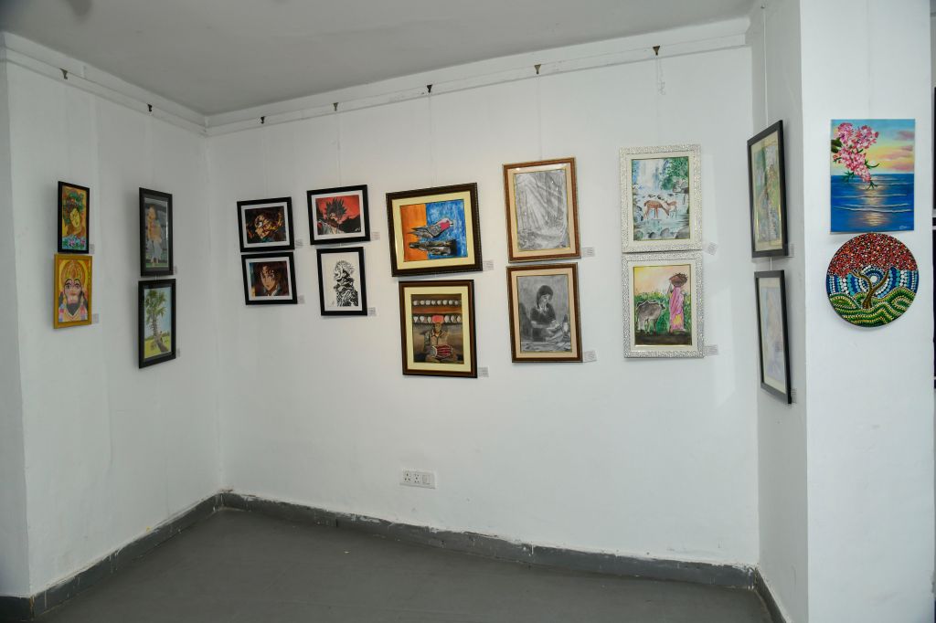 An art exhibit showing various framed artworks on white walls, including paintings and drawings in different styles and colors.