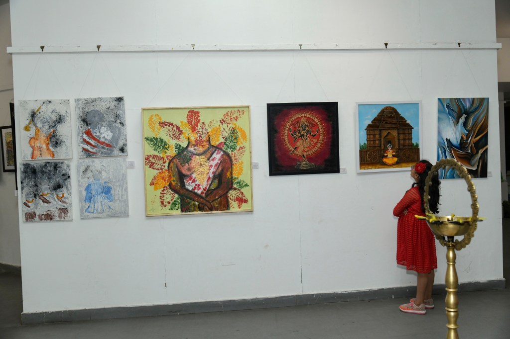 A young girl in a red dress gazes at a display of colorful artwork hung on a gallery wall, featuring various paintings and an ornamental lamp in the foreground.
