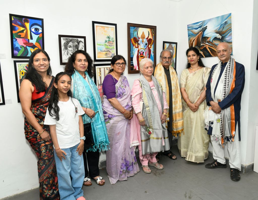A group of seven people posing together in an art gallery, with various colorful artworks displayed on the walls behind them. The group includes women in traditional attire and a young girl, smiling and standing in a line.