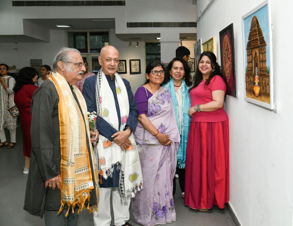 A group of five adults posing together at an art exhibition, showcasing various artworks on the wall behind them. They are dressed in traditional attire, and the setting is a gallery with soft lighting.
