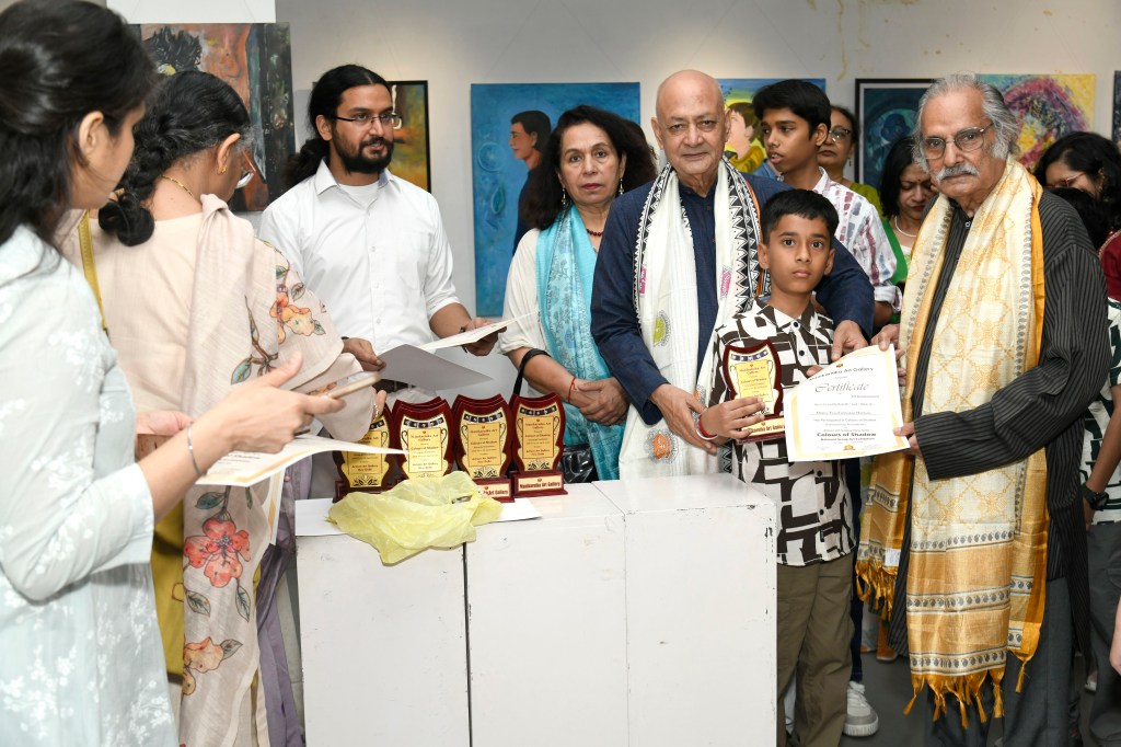 A group of people at an award ceremony, with individuals holding certificates and trophies. A young boy stands in the foreground, displaying an award, surrounded by adults and children in a colorful gallery setting.
