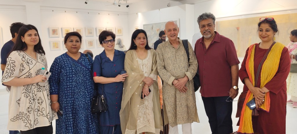 A group of seven individuals posing together in an art gallery, wearing traditional and contemporary clothing, with artworks visible in the background.