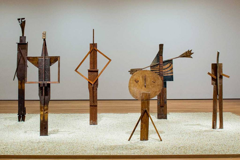 A group of abstract wooden sculptures arranged on a sandy floor in a gallery setting, showcasing various geometric shapes and figures.