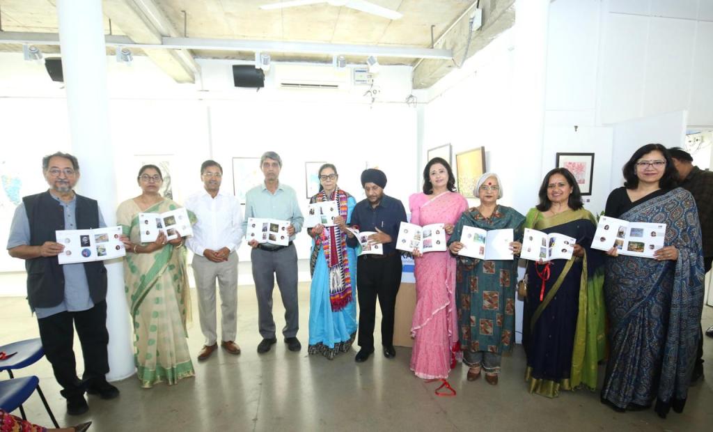 A diverse group of individuals stands together in an art gallery, each holding booklets showcasing various artworks. The group includes men and women dressed in traditional attire, smiling and engaging with the camera.