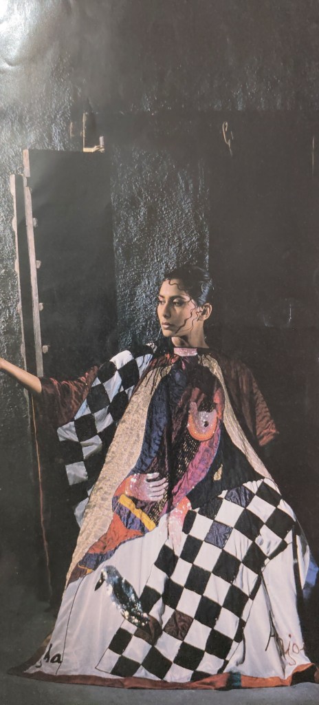 A figure wearing a patterned garment with a checkerboard design and colorful artwork, posed in a dimly lit setting.