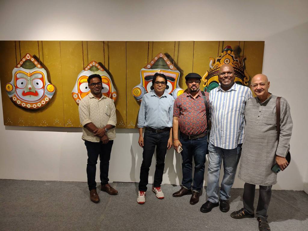 Five men posing in front of colorful traditional art featuring masks on a beige wall. The setting appears to be an art gallery.