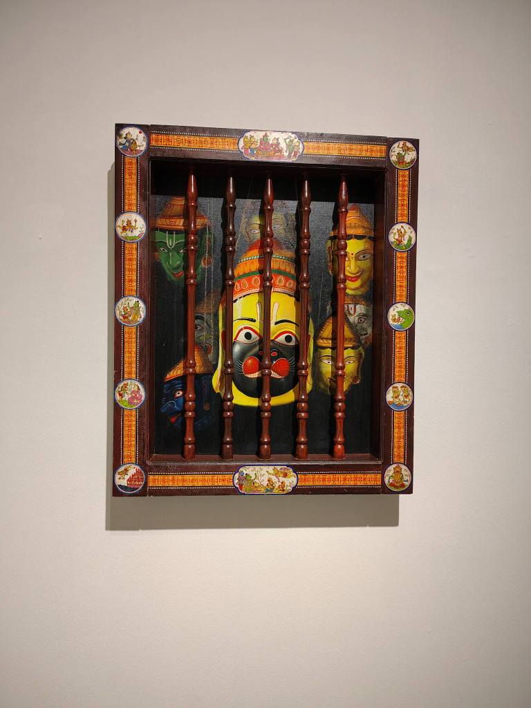 A decorative wall-mounted frame containing colorful traditional masks, separated by wooden bars.