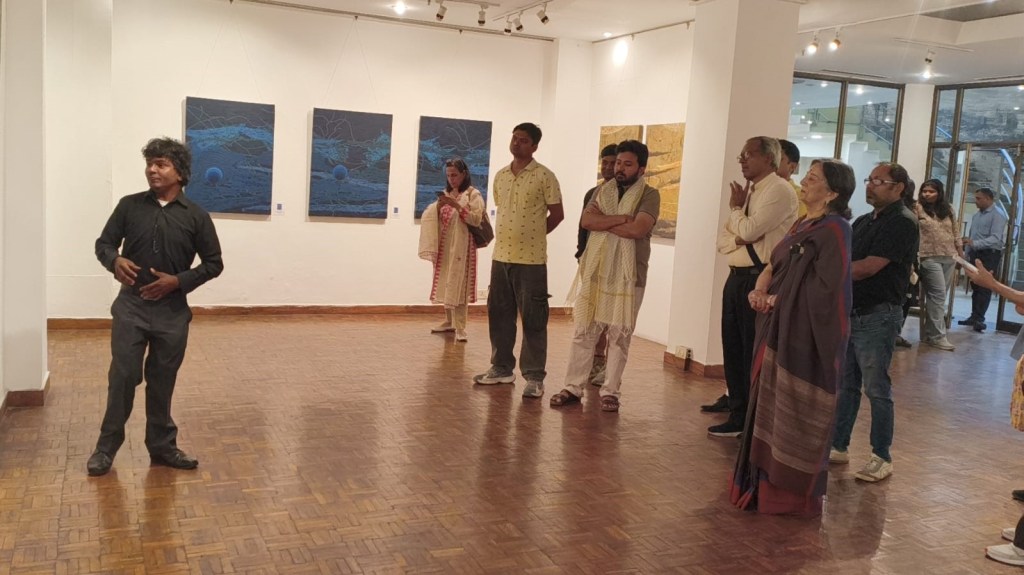 A speaker presenting in an art gallery with audience members listening, in front of blue and gold artwork displayed on the walls.
