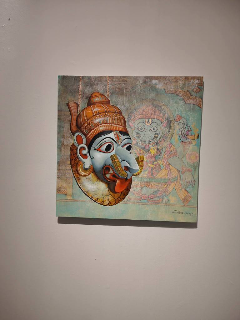 A vibrant artwork featuring a stylized mask of Lord Ganesha, intricately designed with orange and blue tones, mounted against a textured background with faint depictions of other deities.