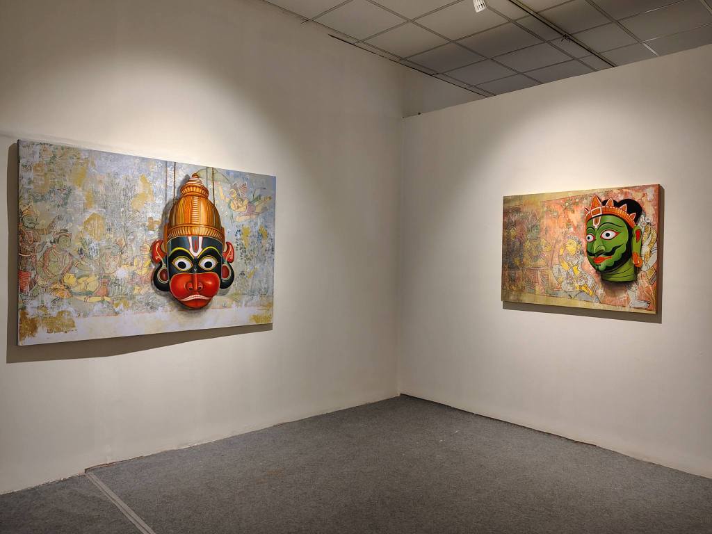 A gallery interior featuring two large paintings of traditional masks on white walls, one with a red and yellow mask, and another with a green mask against a textured background.