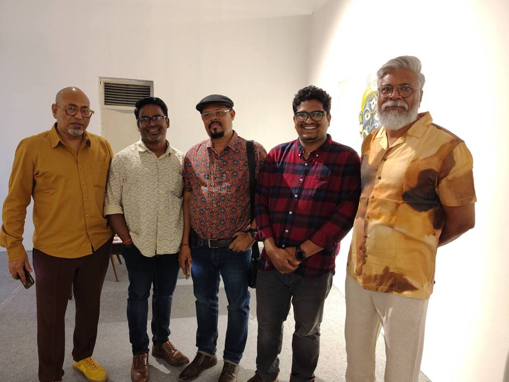 A group of five men standing together in an art gallery, each wearing distinct, stylish clothing. They are smiling and posing for the photo against a light-colored wall.