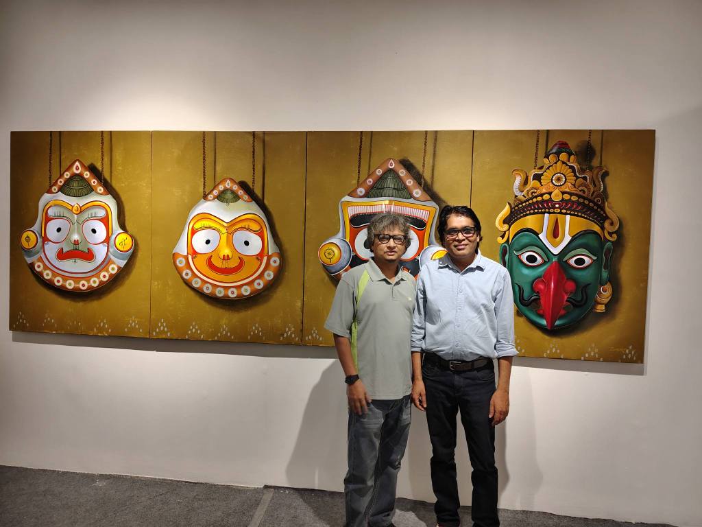 Two individuals stand in front of a large artwork featuring colorful traditional masks on a golden background.
