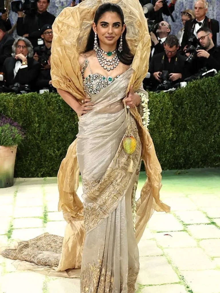 A woman dressed in an intricate silver and gold saree with a lavishly draped golden shawl, adorned with elaborate jewelry, walking confidently on a red carpet at a high-profile event.