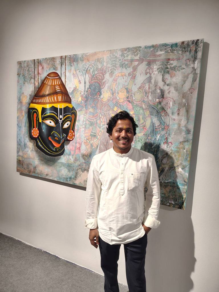 A man standing in front of a colorful artwork depicting a traditional Indian mask and intricate illustrations.