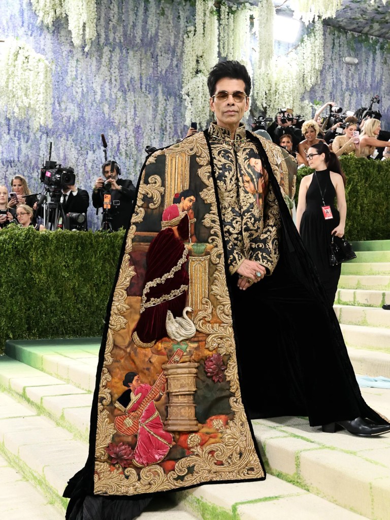 A man wearing an intricately embroidered black cape with colorful scenes, posing confidently on the red carpet surrounded by photographers and event attendees.