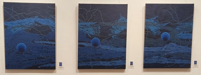 Three abstract paintings featuring deep blue tones, depicting circular shapes and textured patterns, displayed in a gallery setting.