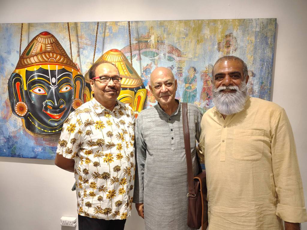 Three men pose for a photo in front of a colorful wall painting featuring traditional Indian art and vibrant masks.