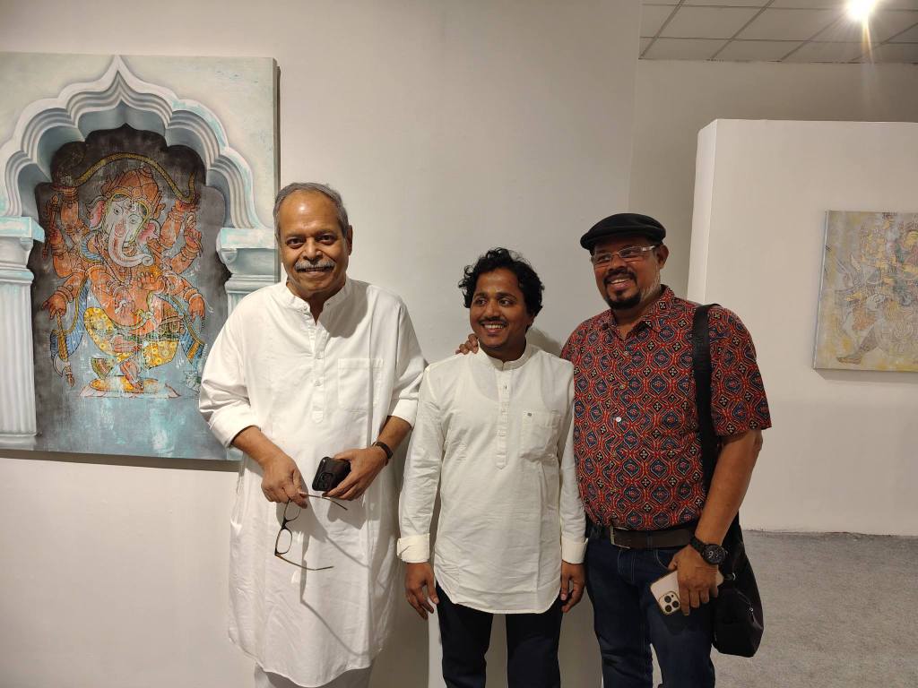 Three men pose together in an art gallery, with a colorful painting of Ganesh visible in the background.