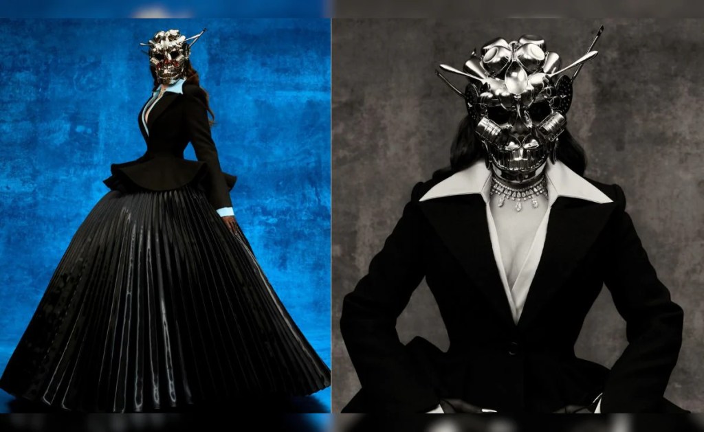 A person wearing a dark, elegant outfit featuring a large, pleated skirt and a fitted jacket, adorned with a unique metallic skull mask decorated with various objects, standing against a colorful abstract background.