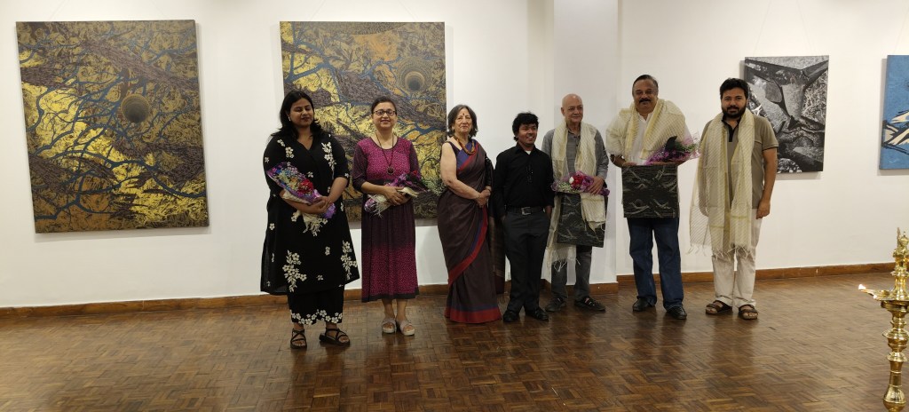 A group of seven individuals standing together in an art gallery, each holding floral arrangements. They are positioned in front of large, colorful paintings. The setting is well-lit with wooden flooring.