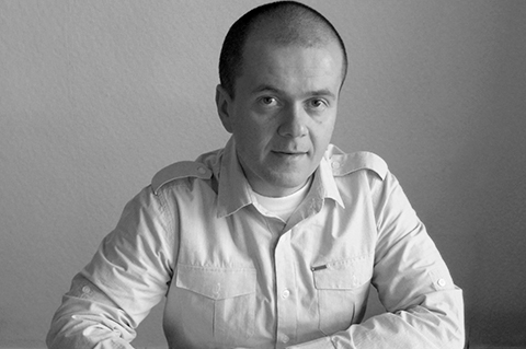A black and white portrait of a man seated at a table, wearing a light-colored button-up shirt and looking directly at the camera.