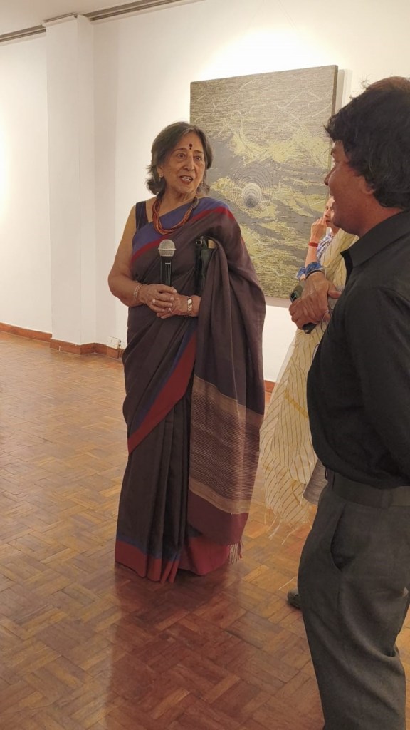 A woman wearing a traditional sari is speaking into a microphone at an art gallery event, while engaged with two individuals nearby.